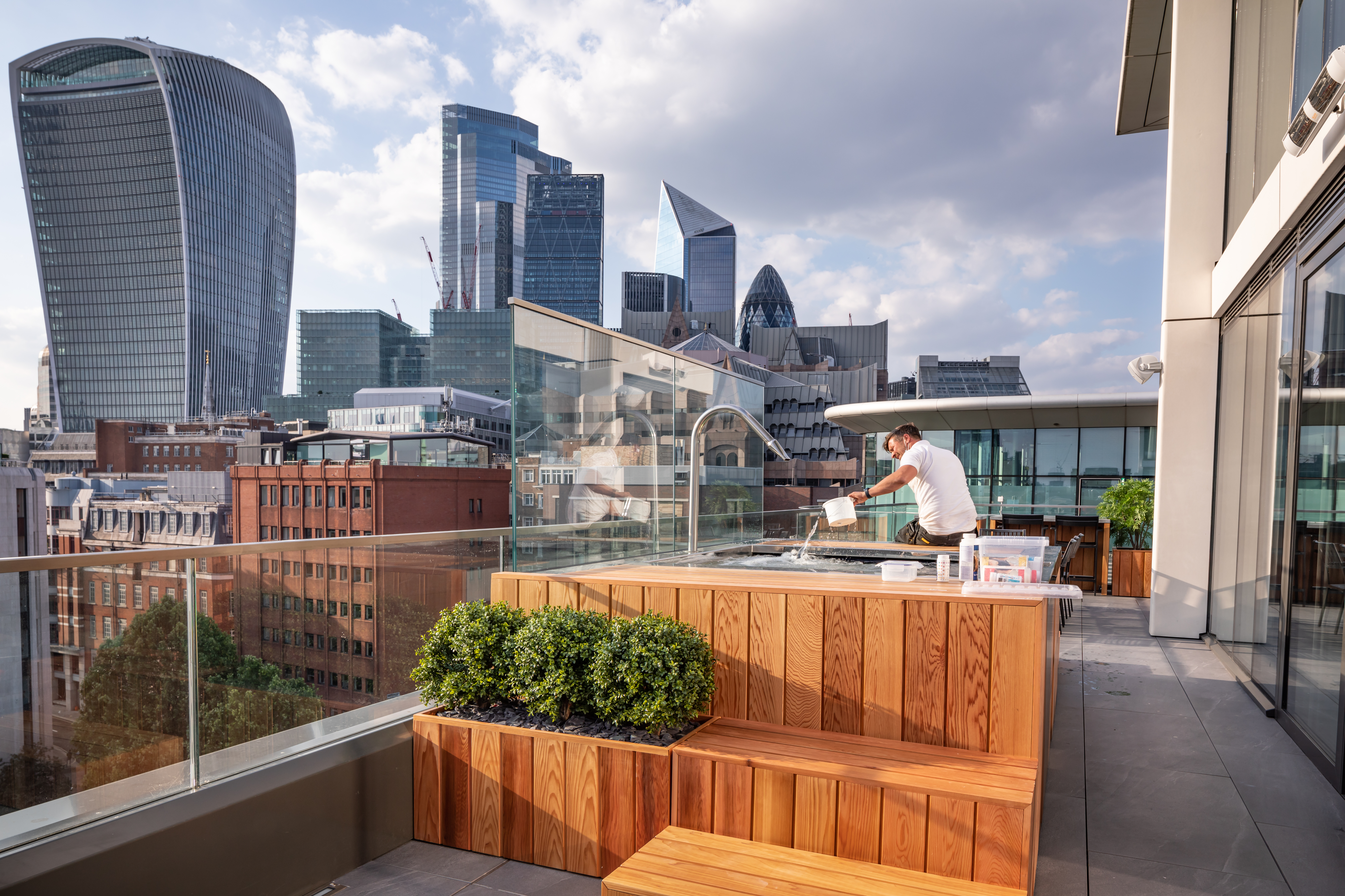 A man in white T Shirt sitting on a Hot Tub as he is serving it for his client.  The hot tub is on a Penthouse Suite Balcony overlooking the Thames and magnificent architecture including the shard and tallest building in London.  Branding Photography for SpaFlo by Di Jones Photography 