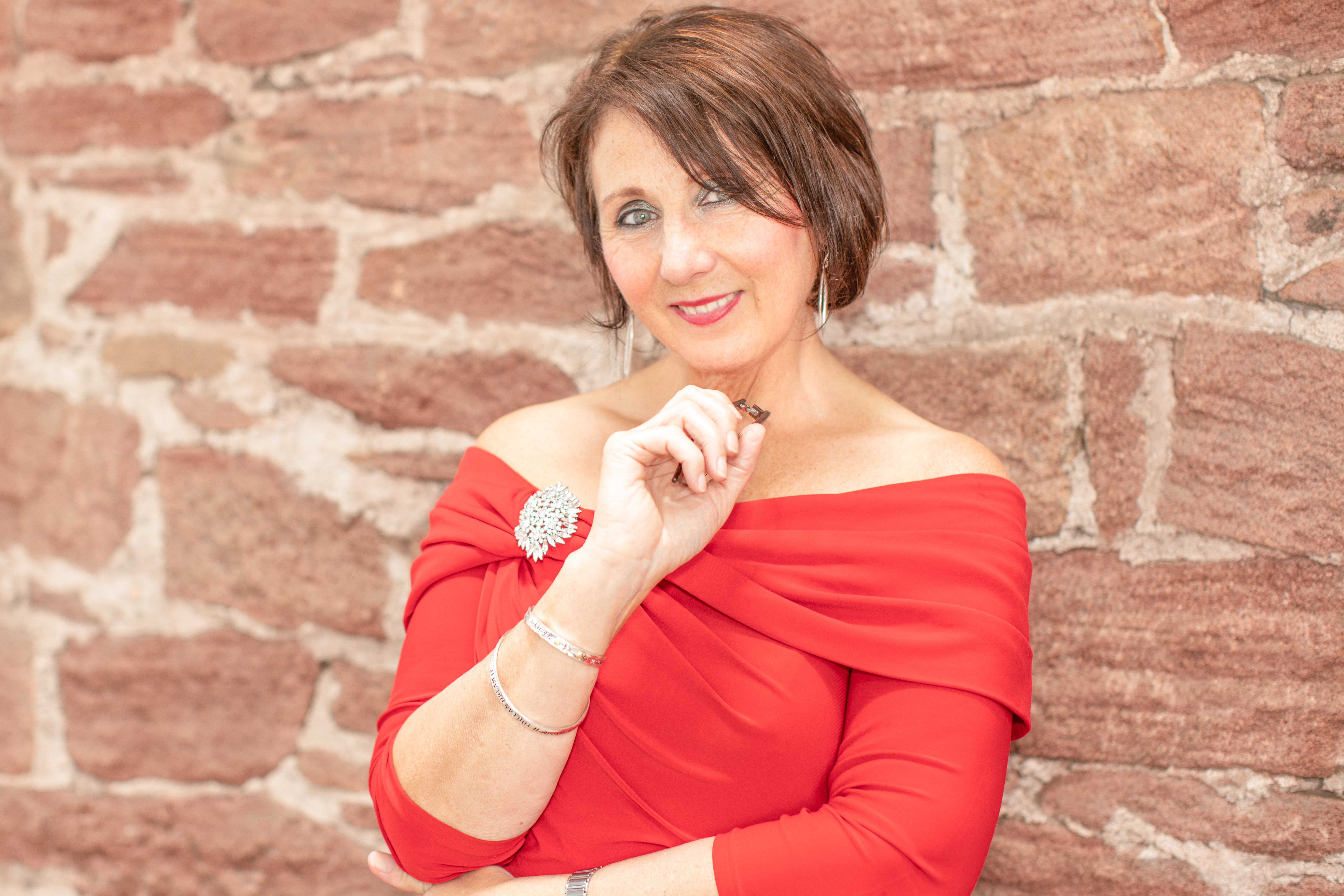 Lady with chin length brown hair, bright green eyes and smiling as she standing next to an internal stone wall, wearing a red dress.  One arm is folded on her waist whilst the other is folded up to her chin holding a pair of reading glasses.  Branding Photography by Di Jones Photography