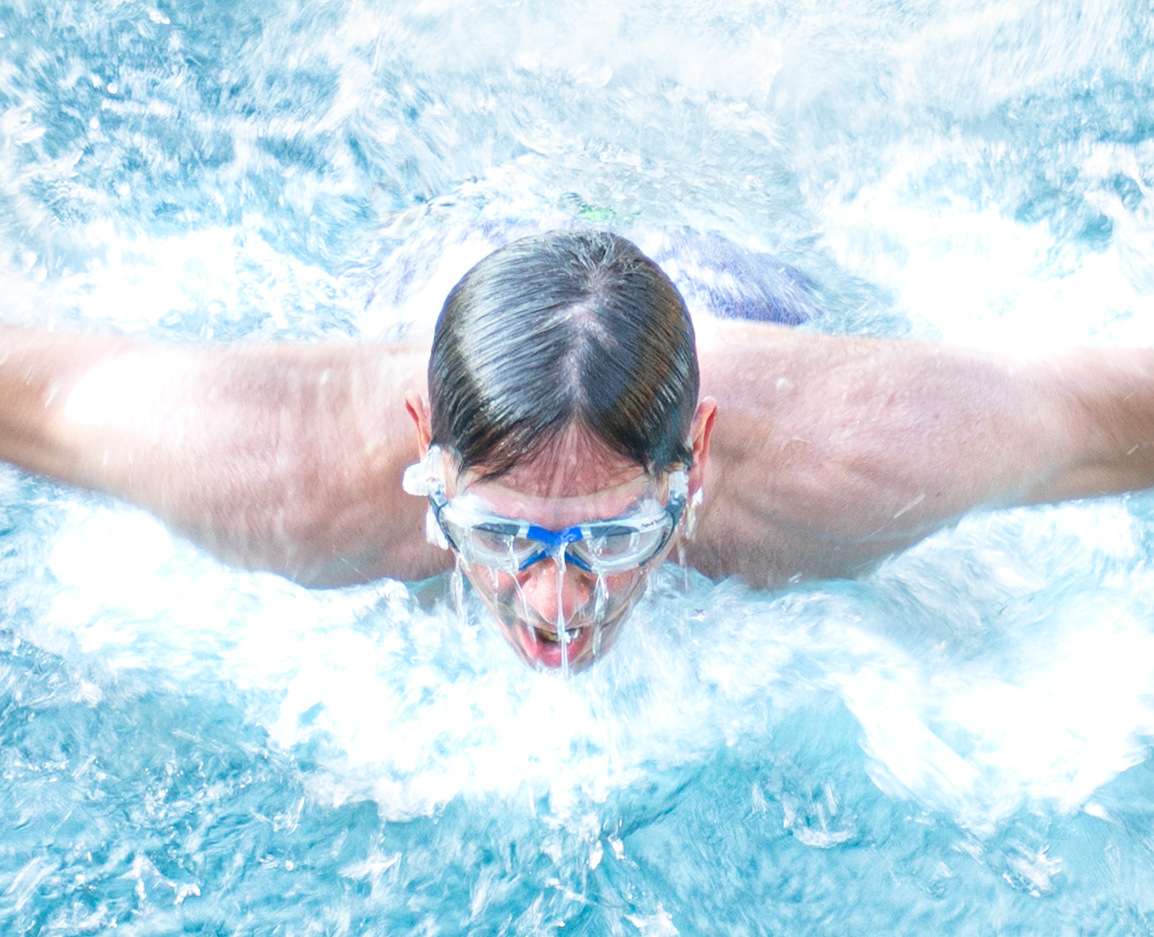 Man wearing swimming goggles with water pouring down over his face as his head rises out of the water, mouth open to catch a breath.  Both arms are stretched wide as he practices his butterfly stroke.  Branding Photography for SpaFlo by Di Jones Photography